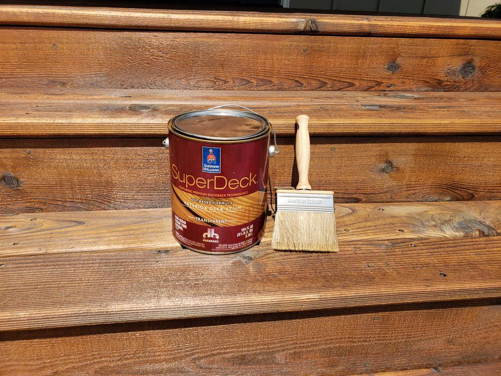 A can of superdeck stain next to a paintbrush on a wooden surface.